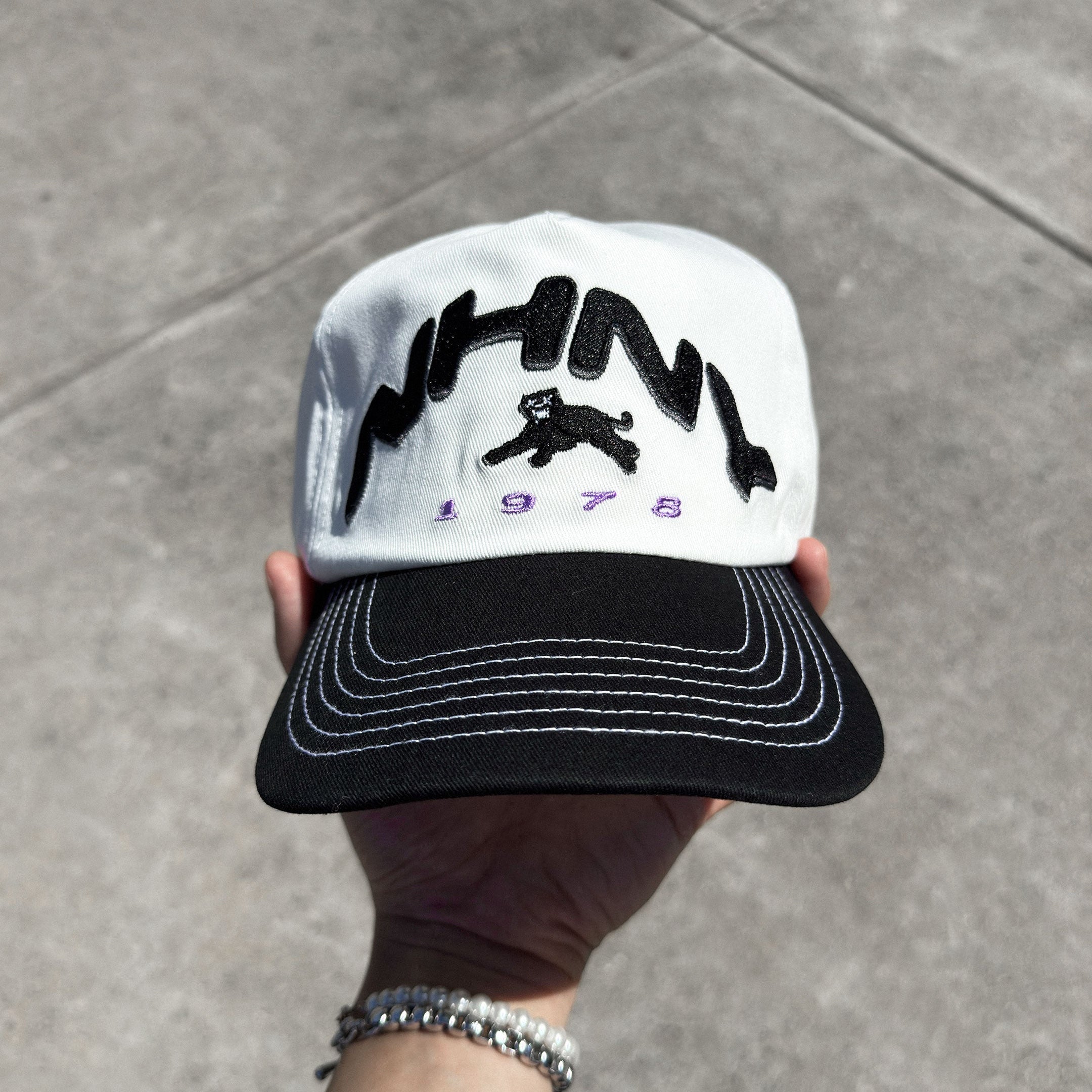 A hand holding a vintage style cap with white brim stitching, black crown, and an embroidered logo on the front.